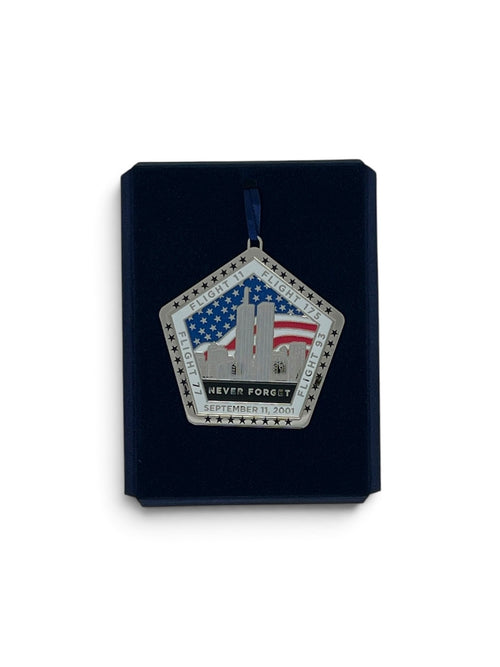 A pentagonal commemorative hanging ornament displayed on a dark navy presentation card against a white background. The ornament features the Twin Towers, an American flag, stars, and the text 'NEVER FORGET' and 'SEPTEMBER 11, 2001'.