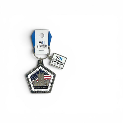 A 9/11 Memorial & Museum keychain featuring a pentagonal charm with the Twin Towers, American flag, and 'Never Forget' text, along with a small square tag and blue-and-white retail hanger, shown on a white background.