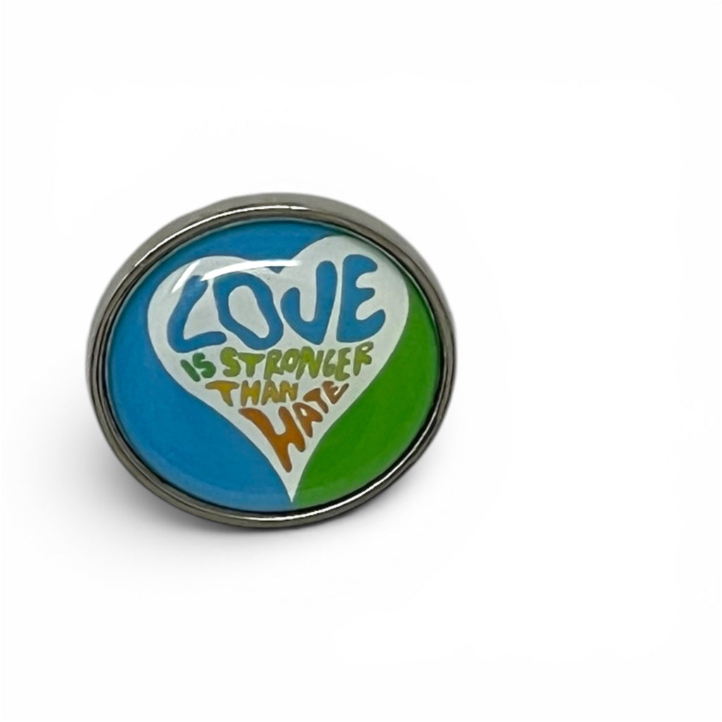 A round metal pin featuring a colorful heart design with the text 'LOVE IS STRONGER THAN HATE' in blue, green, white, and orange.