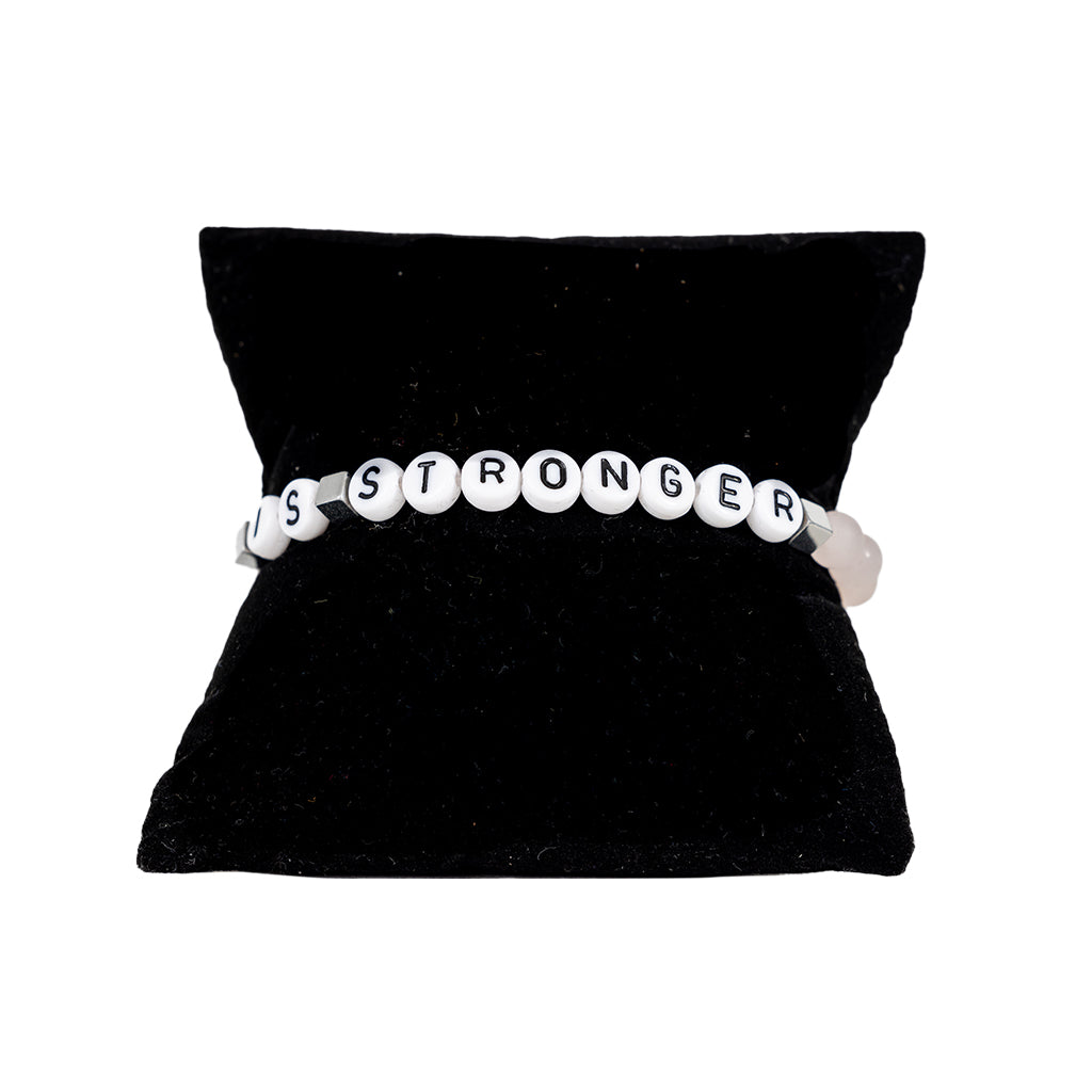 A pale pink elastic stretch bracelet with white text 'LOVE IS STRONGER', presented on a black cushion display.