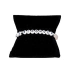 A pale pink elastic stretch bracelet with white text 'LOVE IS STRONGER', presented on a black cushion display.