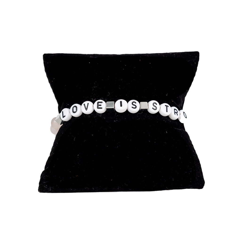 A pale pink elastic stretch bracelet with white text 'LOVE IS STRONGER', presented on a black cushion display.