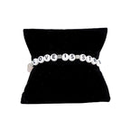 A pale pink elastic stretch bracelet with white text 'LOVE IS STRONGER', presented on a black cushion display.