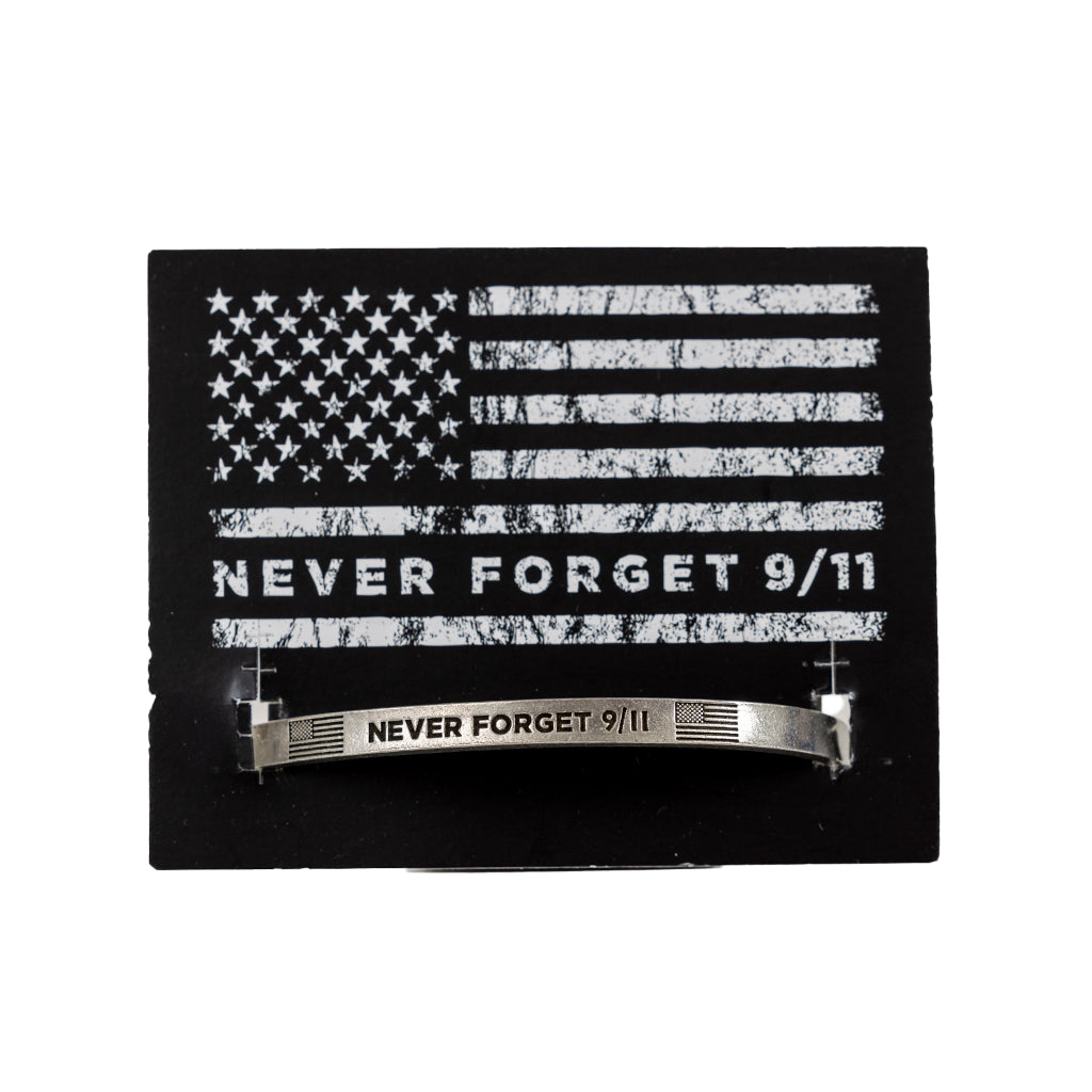 A silver pewter cuff bracelet with the inscription 'NEVER FORGET 9/11' on a black card backer with a tonal American flag and the words 'NEVER FORGET 9/11'.