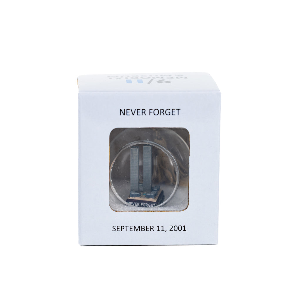 A glass keepsake ornament with a gold-colored cord, containing a miniature replica of the Twin Towers in a small grey square box with the words 'NEVER FORGET' and 'SEPTEMBER 11, 2001'.