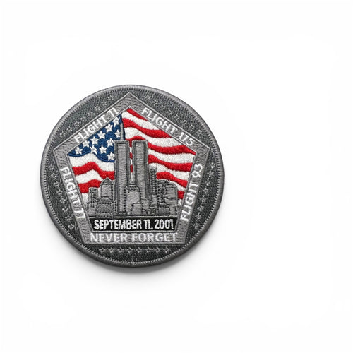 A circular commemorative patch on a plain white background. The design features a gray border with flight numbers, 'SEPT 11, 2001' and 'NEVER FORGET' around an embroidered American flag and city skyline motif.