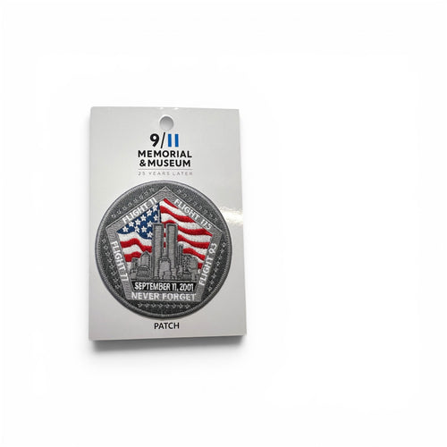 A circular memorial patch mounted on a white backing card. The patch features a silver-toned border with the words 'September 11, 2001' and 'Never Forget,' with an American flag and the New York City skyline in the center.