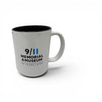 A white ceramic mug with a black interior, featuring the text '9/11 MEMORIAL & MUSEUM 25 YEARS LATER' printed in black and blue, set against a plain white background.