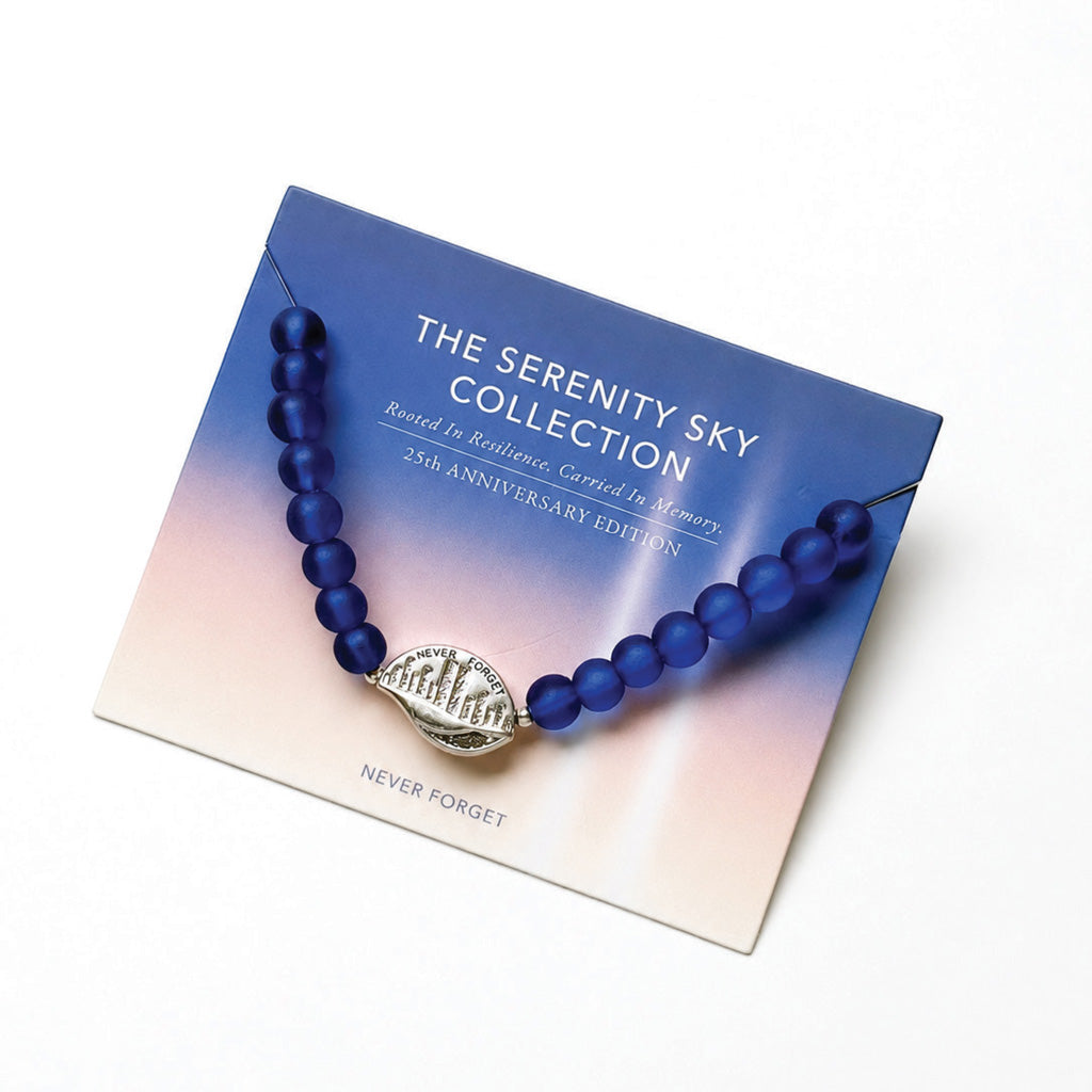 A blue beaded bracelet with a silver-toned oval medallion displaying the NYC skyline, presented on a branded display card reading 'THE SERENITY SKY COLLECTION' and '25th ANNIVERSARY EDITION'.