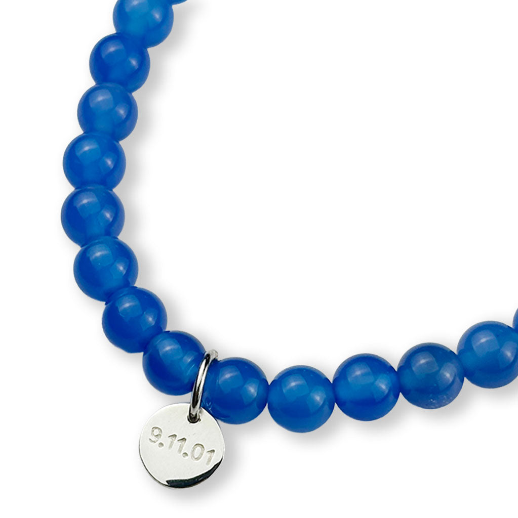 Blue beaded bracelet with a silver distressed piece and a silver charm that says NYC on one side and 9.11.01 on the other, on a white background.