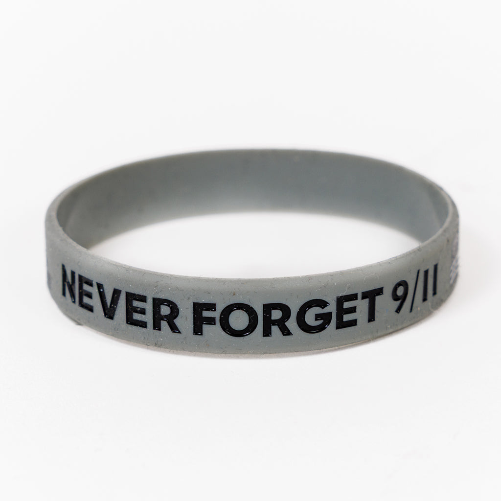 A grey rubber bracelet with the text 'NEVER FORGET 9/11', American Flag and Skyline printed on it.