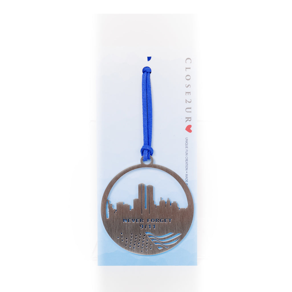 A stainless steel circular keepsake with a skyline silhouette and the text 'NEVER FORGET 9/11' inscribed on it, attached to a blue ribbon on a white to blue ombre card backer.
