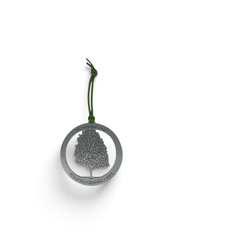 A silver metal keepsake featuring a tree silhouette inside a circular frame with the words RESILIENCE • RENEWAL • Hope engraved along the bottom edge, hanging from a dark green cord on a white background.
