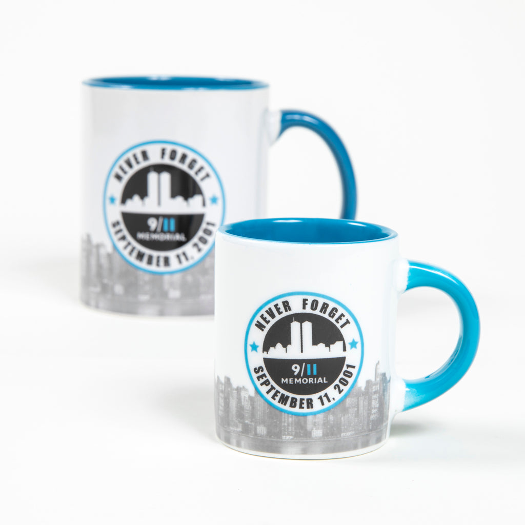 A white mini mug with a light blue interior and handle and a graphic print on the front featuring the Twin Towers silhouette and the text 'Never Forget' 'September 11, 2001 along with the words 9/11 Memorial & Museum logo.