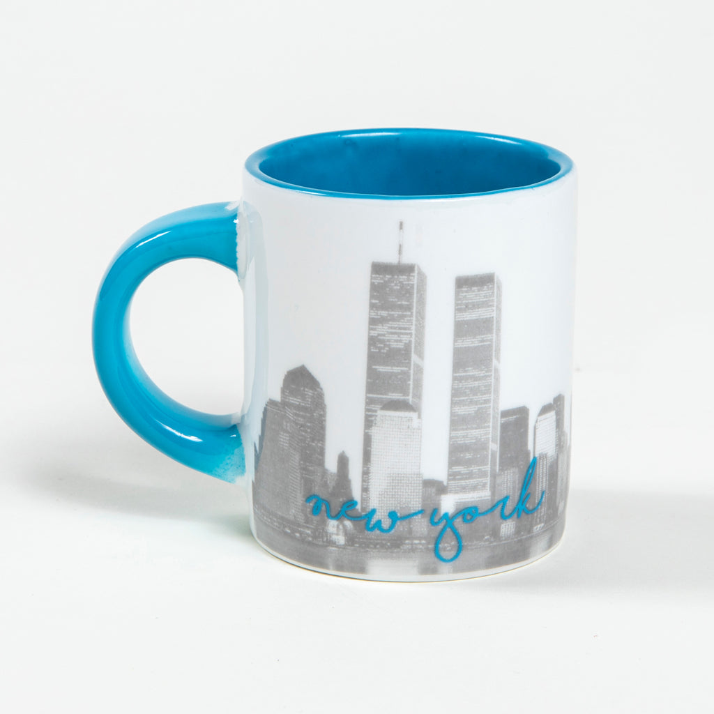 The back view of a white mini mug with a light blue interior and handle and a graphic print on the front featuring the Twin Towers silhouette and the text 'Never Forget' 'September 11, 2001 along with the words 9/11 Memorial & Museum logo.