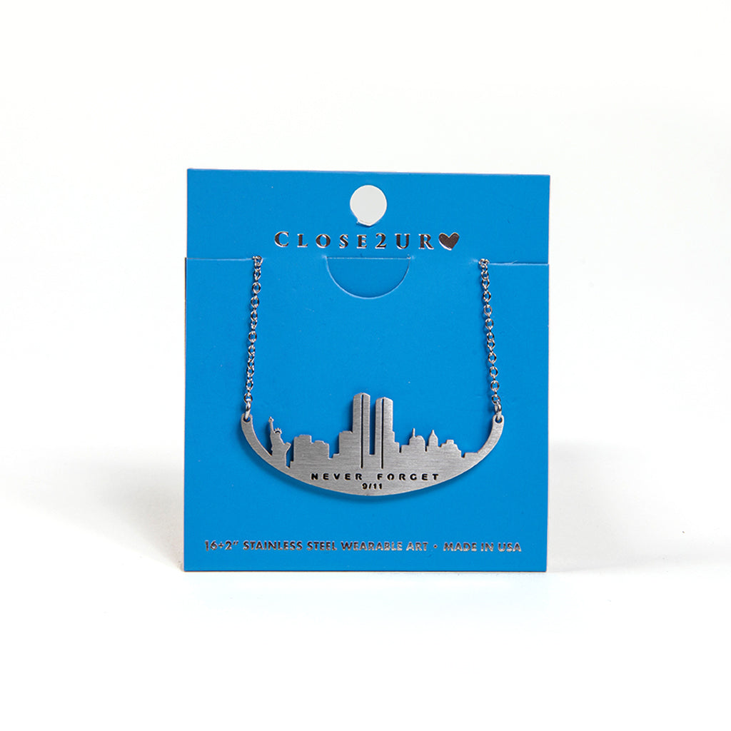 A stainless steel necklace with a skyline silhouette and the text 'NEVER FORGET 9/11' inscribed on it, on a blue card backer.