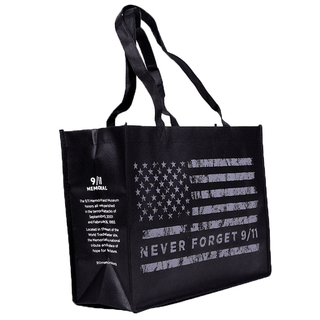 A black tote bag with a grey American flag design and the text 'NEVER FORGET 9/11' on it with the 9/11 Memorial logo and mission-based text on the side gusset.